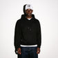 A man wearing a black pullover COMUNE Luka hoodie with a small tonal embroidered logo on the left chest, a white baseball cap with black embroidery lettering, and dark blue jeans, standing against a plain white background.