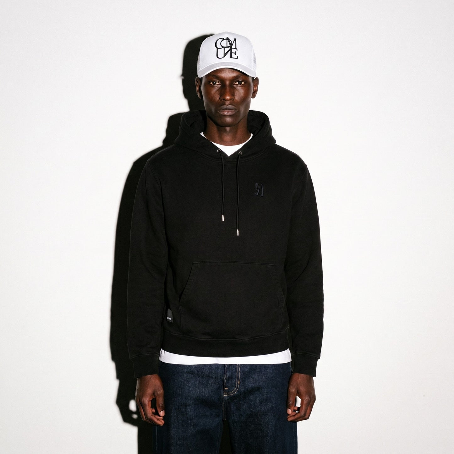 A man wearing a black pullover COMUNE Luka hoodie with a small tonal embroidered logo on the left chest, a white baseball cap with black embroidery lettering, and dark blue jeans, standing against a plain white background.