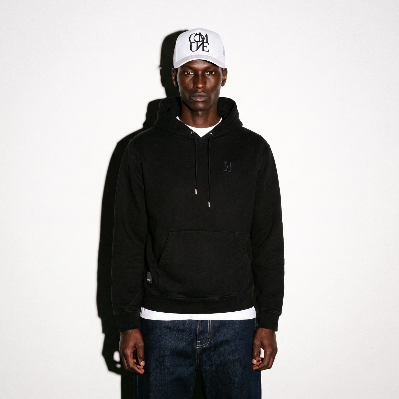 A man wearing a black pullover COMUNE Luka hoodie with a small tonal embroidered logo on the left chest, a white baseball cap with black embroidery lettering, and dark blue jeans, standing against a plain white background.
