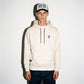 A man wearing a bone color wash COMUNE Luka hoodie with a small black logo on the left chest, a matching black and white cap, and dark blue jeans, standing against a white wall.