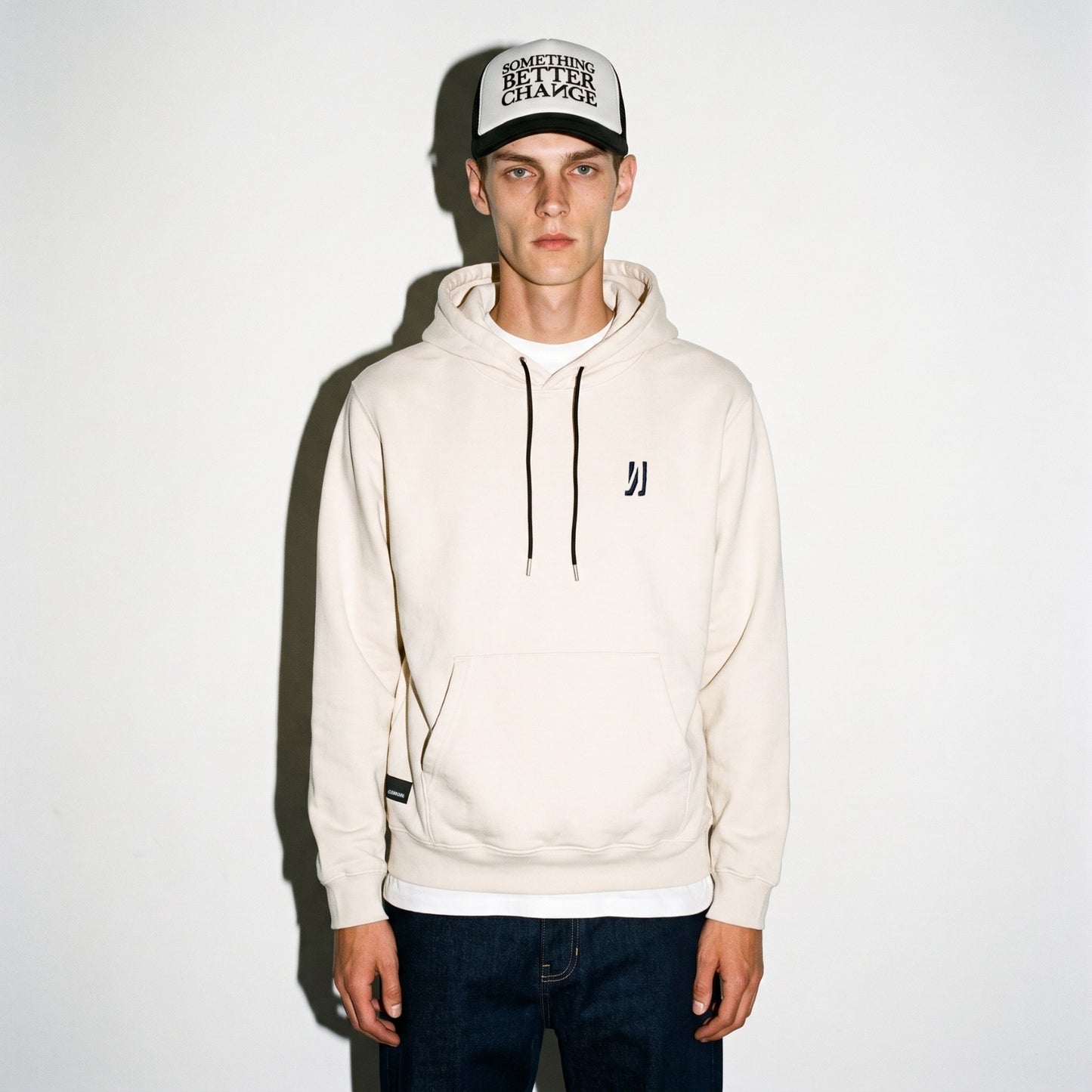 A man wearing a bone color wash COMUNE Luka hoodie with a small black logo on the left chest, a matching black and white cap, and dark blue jeans, standing against a white wall.