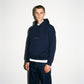 A man wearing a dark navy blue COMUNE Luka pullover hoodie with a small embroidered logo on the left chest, paired with dark jeans and a white undershirt, standing against a plain off-white background.