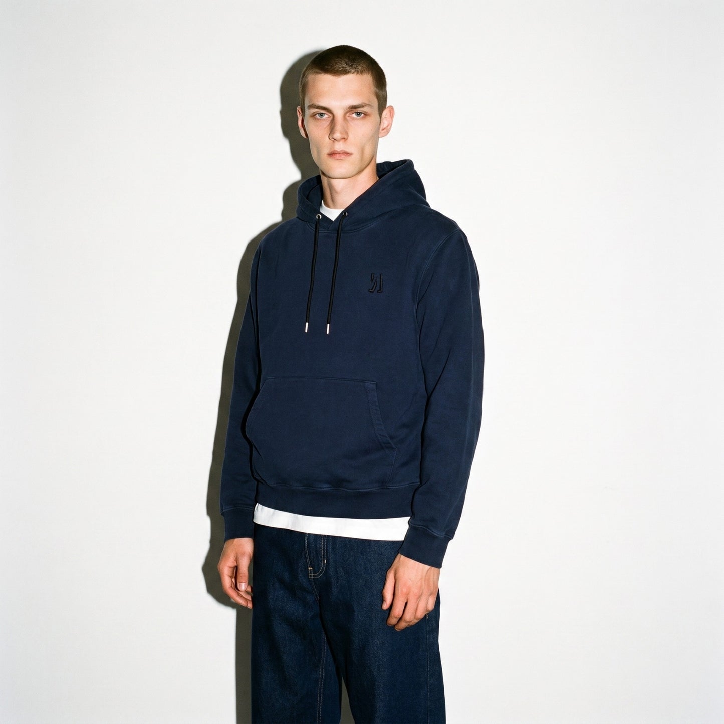A man wearing a dark navy blue COMUNE Luka pullover hoodie with a small embroidered logo on the left chest, paired with dark jeans and a white undershirt, standing against a plain off-white background.