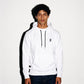 A man wearing a white COMUNE Luka pullover hoodie with black drawstrings and a small black reversed 'N' logo on the left chest, paired with dark blue jeans, standing against a white wall with a hard shadow.