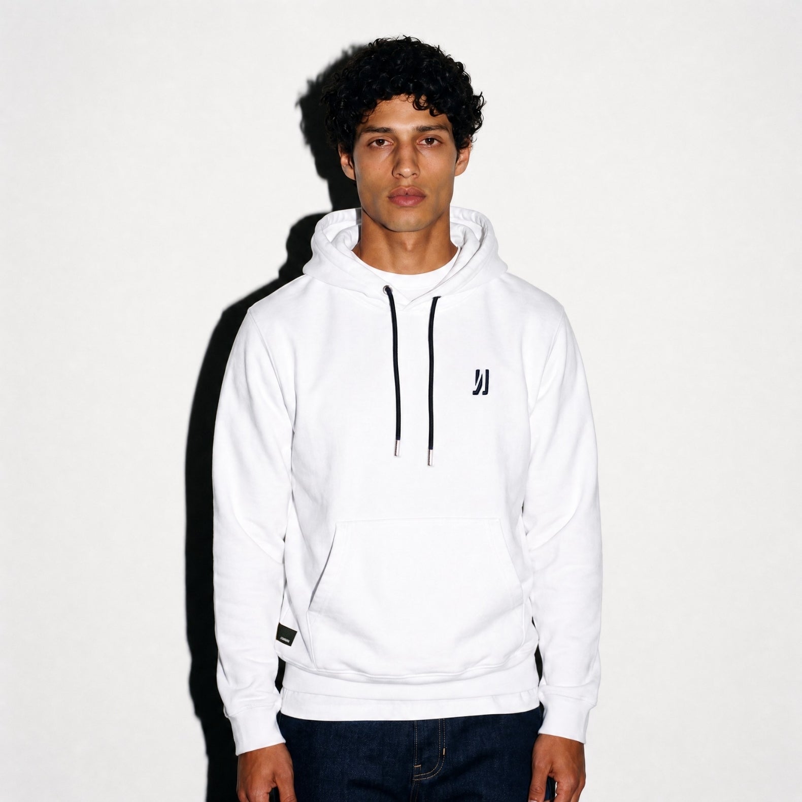 A man wearing a white COMUNE Luka pullover hoodie with black drawstrings and a small black reversed 'N' logo on the left chest, paired with dark blue jeans, standing against a white wall with a hard shadow.