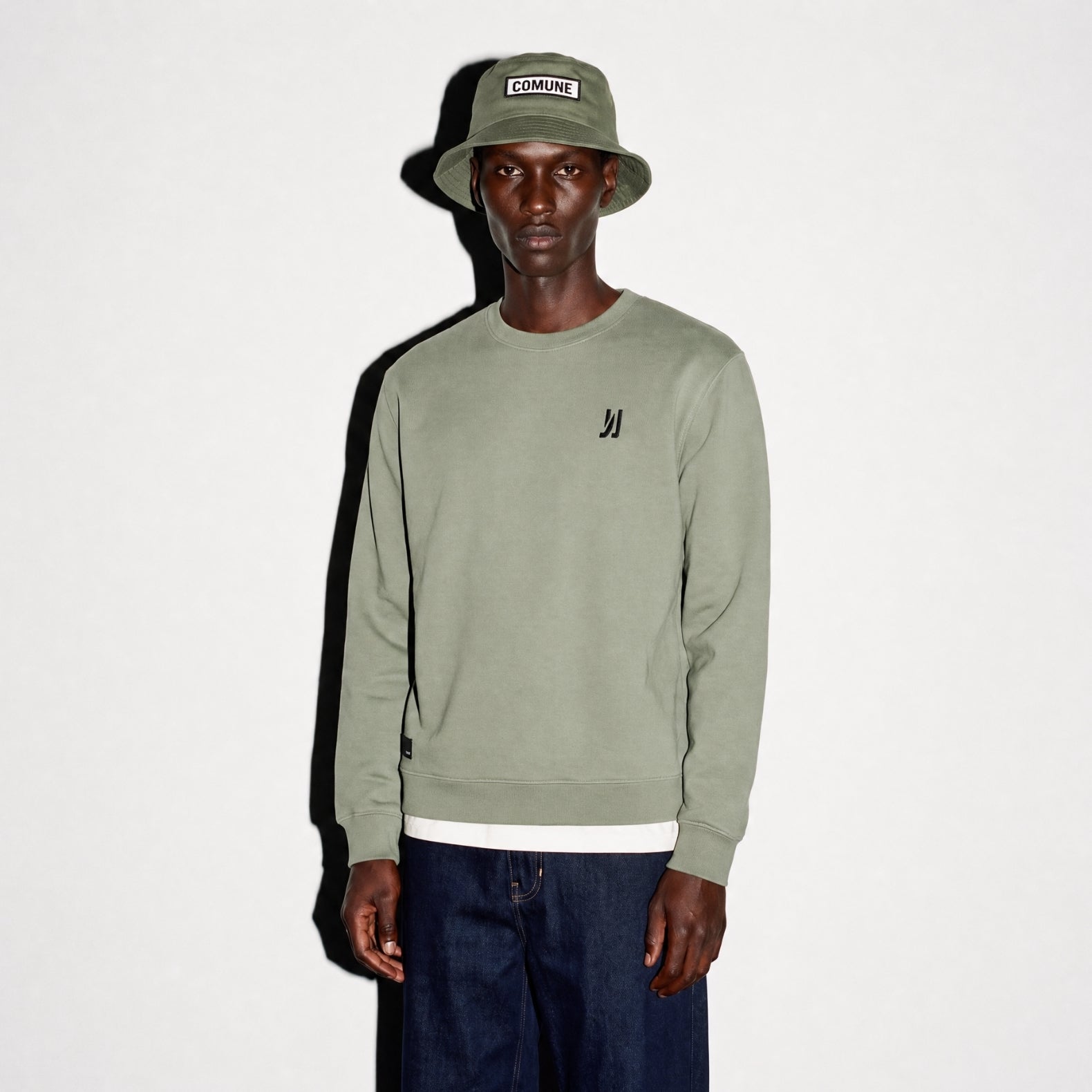 A model wearing a sage color COMUNE Luka Crew sweatshirt with a small black embroidered logo on the left chest, paired with a matching army green bucket hat and dark blue jeans, standing against a plain white background.