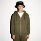 A model wearing an army green COMUNE Luka Zip-up hoodie and matching sweatpants with a black bucket hat featuring the word 'Comune' in white, standing against a plain white background.