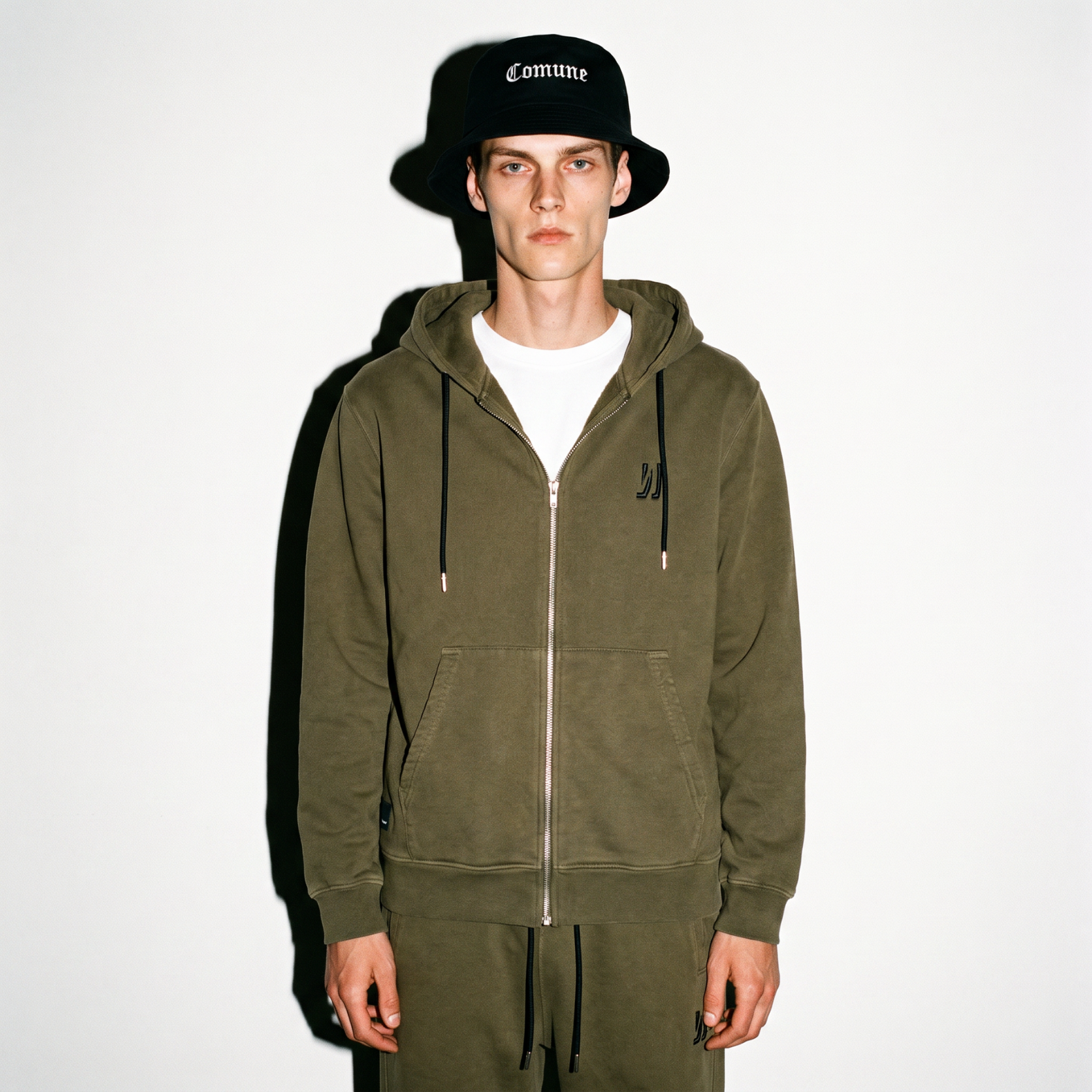 A model wearing an army green COMUNE Luka Zip-up hoodie and matching sweatpants with a black bucket hat featuring the word 'Comune' in white, standing against a plain white background.