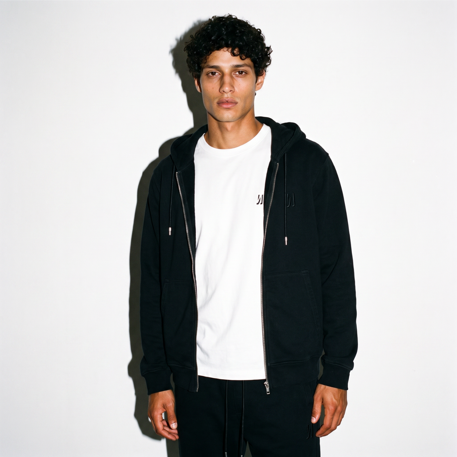 A man wearing a black COMUNE Luka zip-up hoodie over a white t-shirt and black pants, standing against a plain white background with a strong shadow.