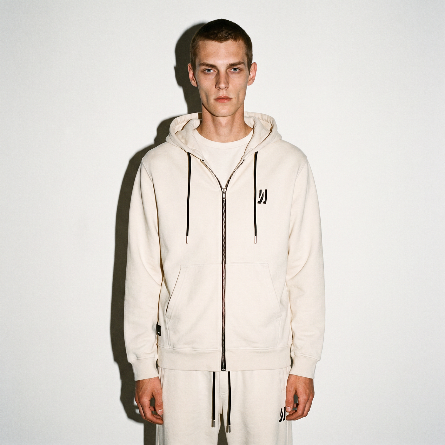 A model wearing a bone color washed COMUNE Luka Zip-up hoodie and matching sweatpants, standing against a plain white background.