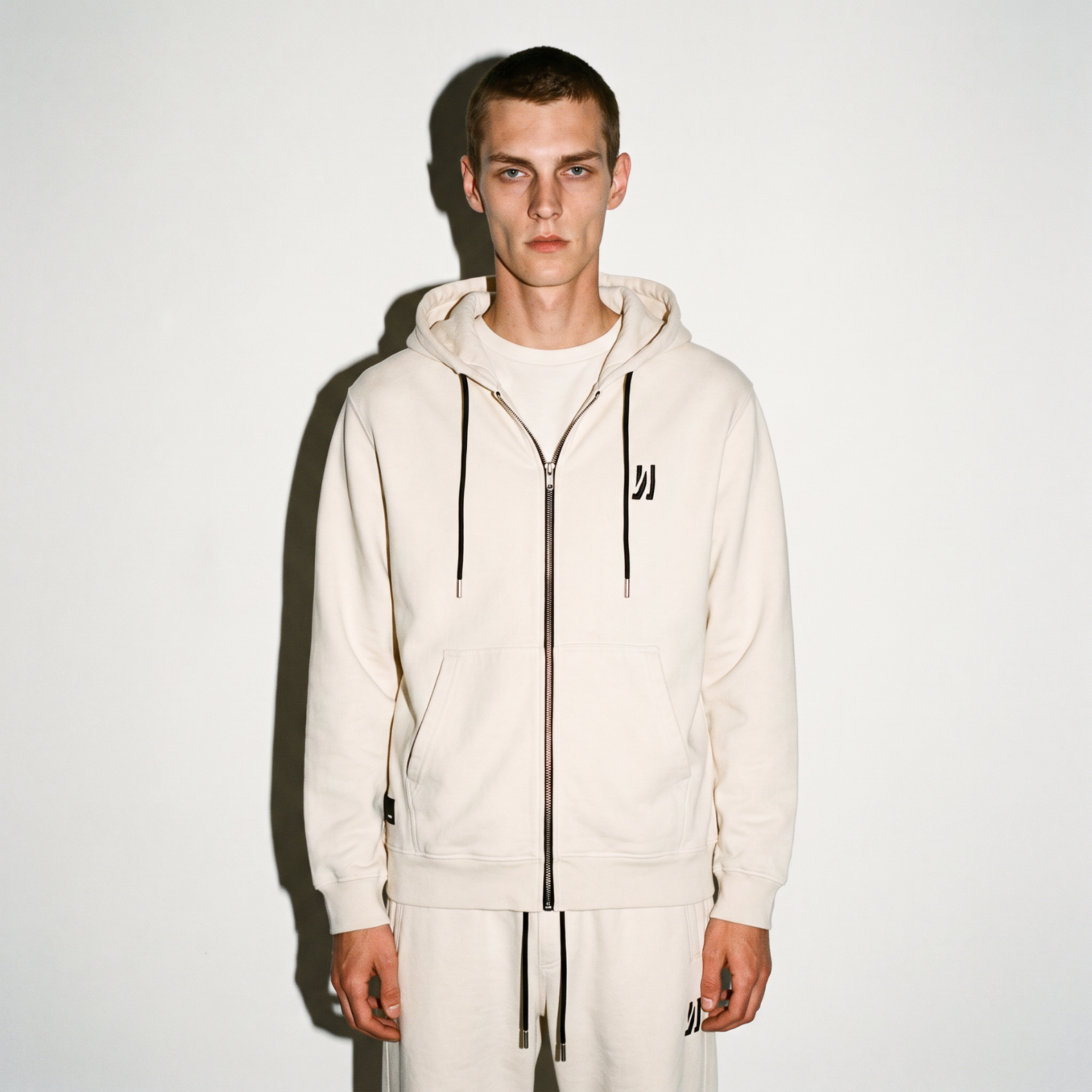 A model wearing a bone color washed COMUNE Luka Zip-up hoodie and matching sweatpants, standing against a plain white background.