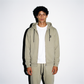 A model wearing a sage color washed COMUNE Luka Zip-up hoodie and matching sweatpants with white t-shirt, standing against a plain white background.