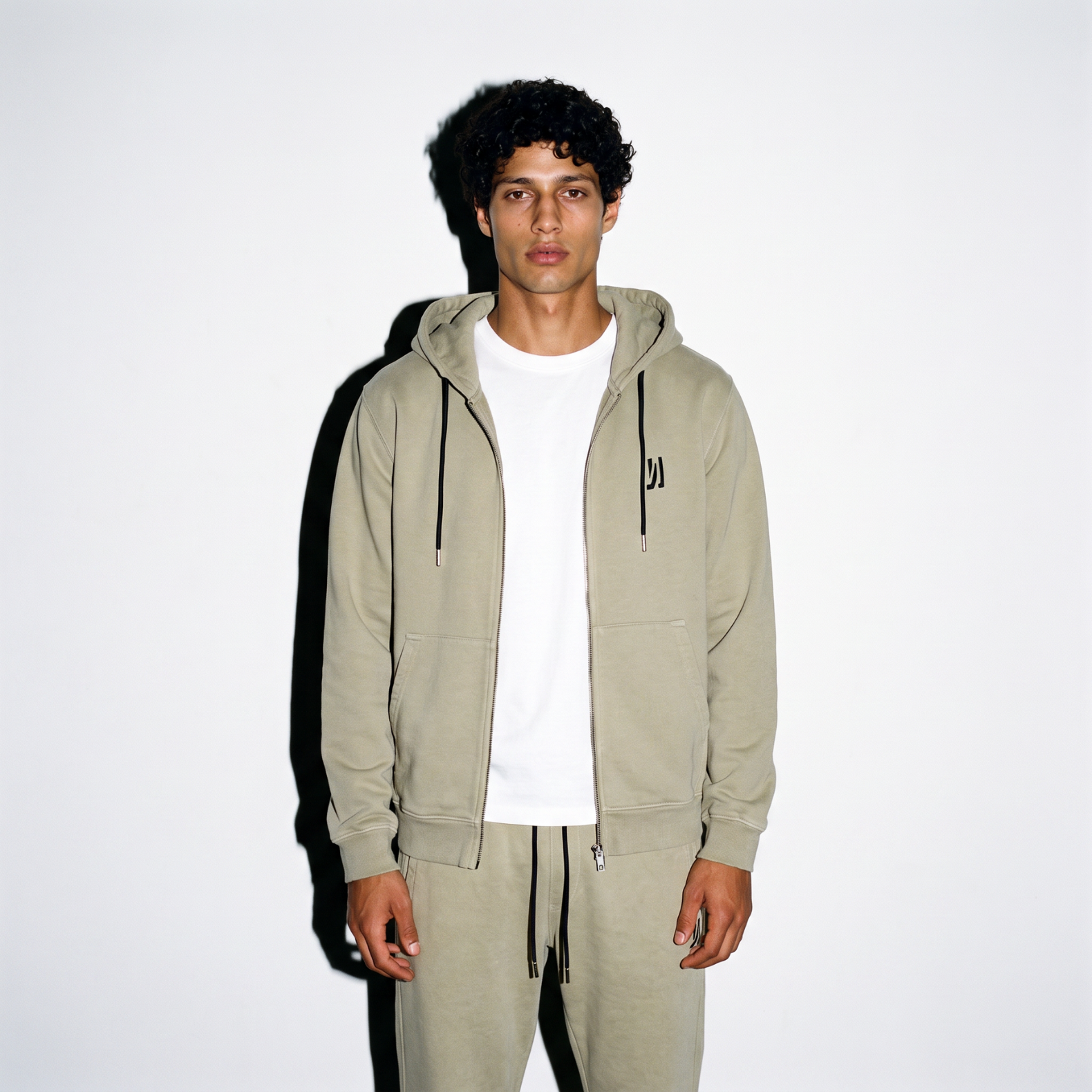A model wearing a sage color washed COMUNE Luka Zip-up hoodie and matching sweatpants with white t-shirt, standing against a plain white background.
