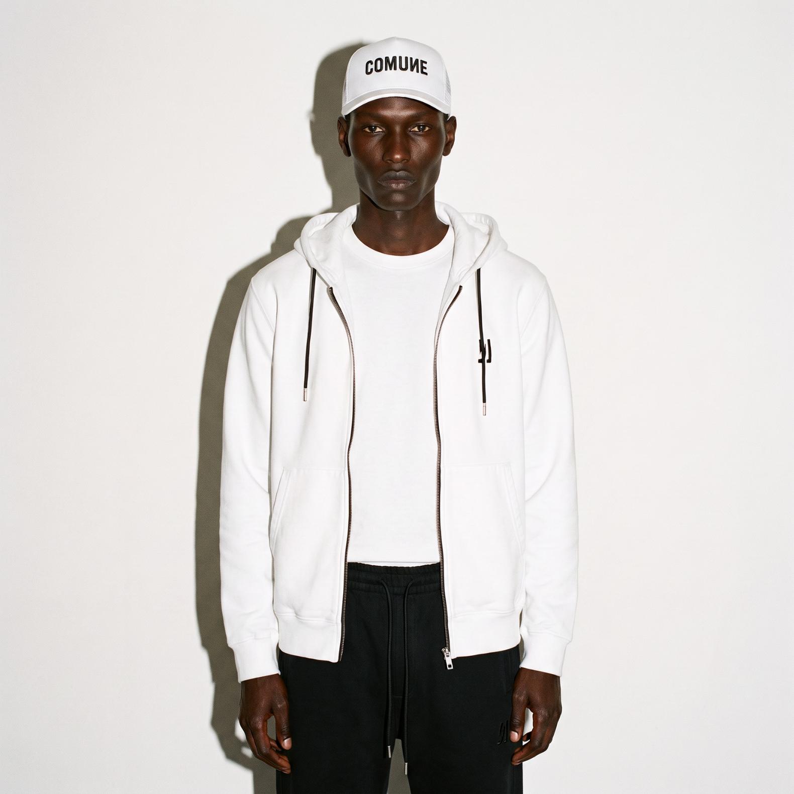 A model wearing a white COMUNE Luka Zip-up hoodie and matching sweatpants with a white trucker hat featuring the word 'Comune' in black embroidery, standing against a plain white background.