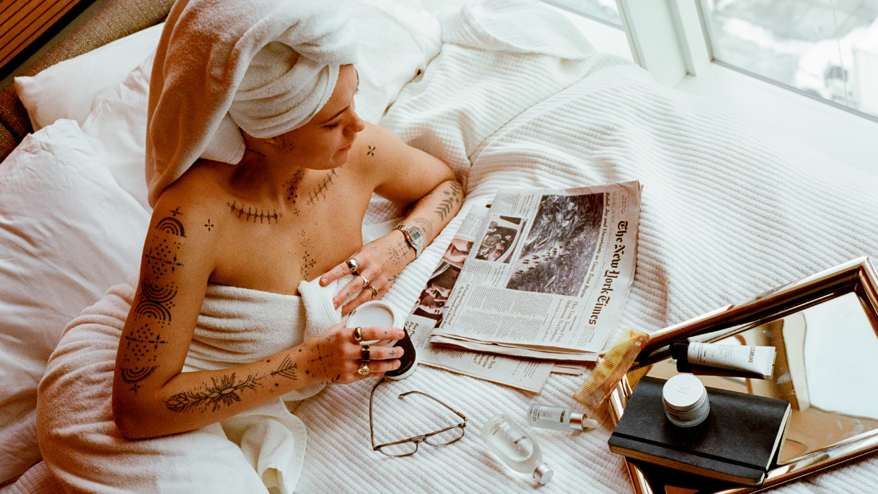 Comune model laying on the bed reading newspaper and Comune Essentials Skincare products laying around.