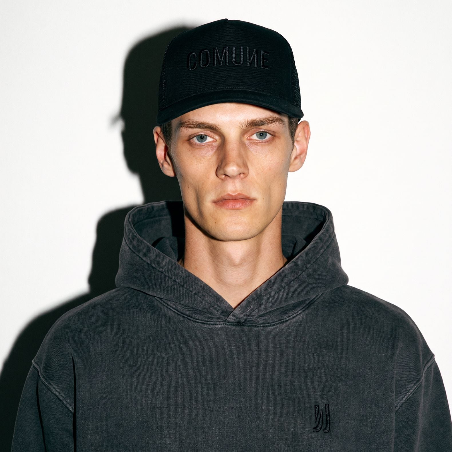 A man wearing a black COMUNE Tonal trucker hat with 'COMIUNE' embroidered on the front and a dark charcoal gray hooded sweatshirt, photographed against a white background with a strong shadow.