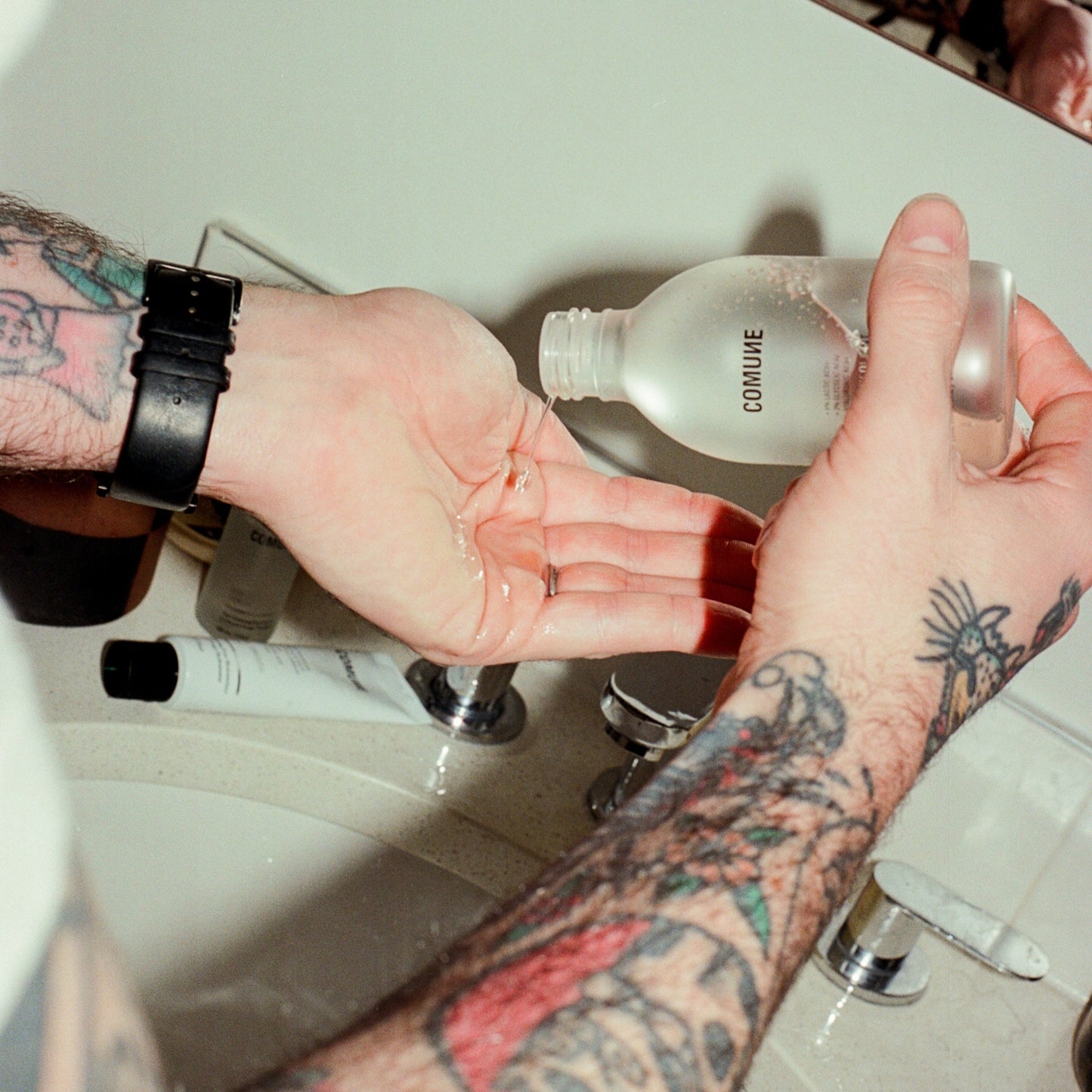 COMUNE artist Ethan with tattoos applying a clear liquid from a bottle Comune AHA Exfoliant Toner to his hand.  Getting ready to apply to his face for brighter, smoother skin result.