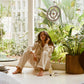 Comune model wearing 'COMUNE' clothes sitting on a floor with plants and a window in the background, holding Comune AHA Exfoliant Toner