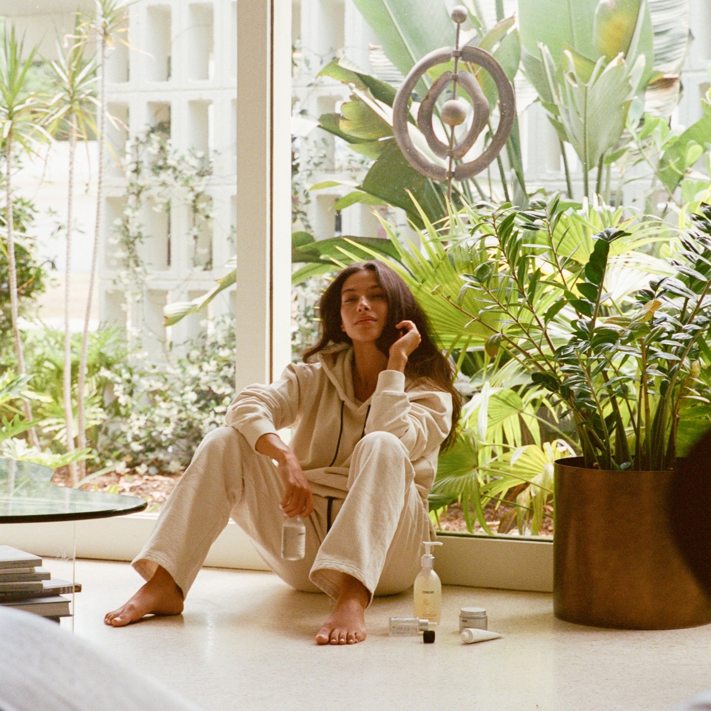 Comune model wearing 'COMUNE' clothes sitting on a floor with plants and a window in the background, holding Comune AHA Exfoliant Toner