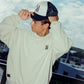 Person wearing a light-colored COMUNE sweatshirt and cap with a scenic background. COMUNE Duke Crew | Bone