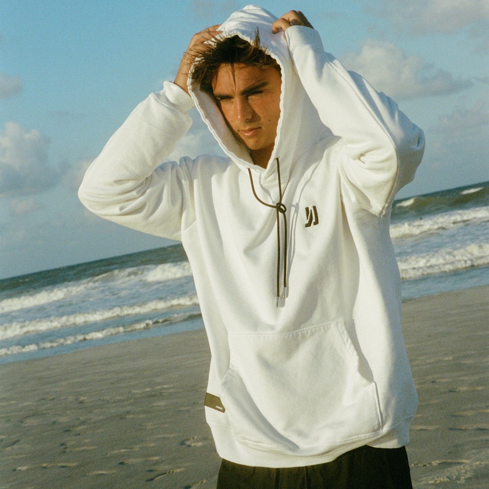 A surfer standing at the beach in front of the waves wearing COMUNE Luka Hoodie. COMUNE LUKA HOODIE | White
