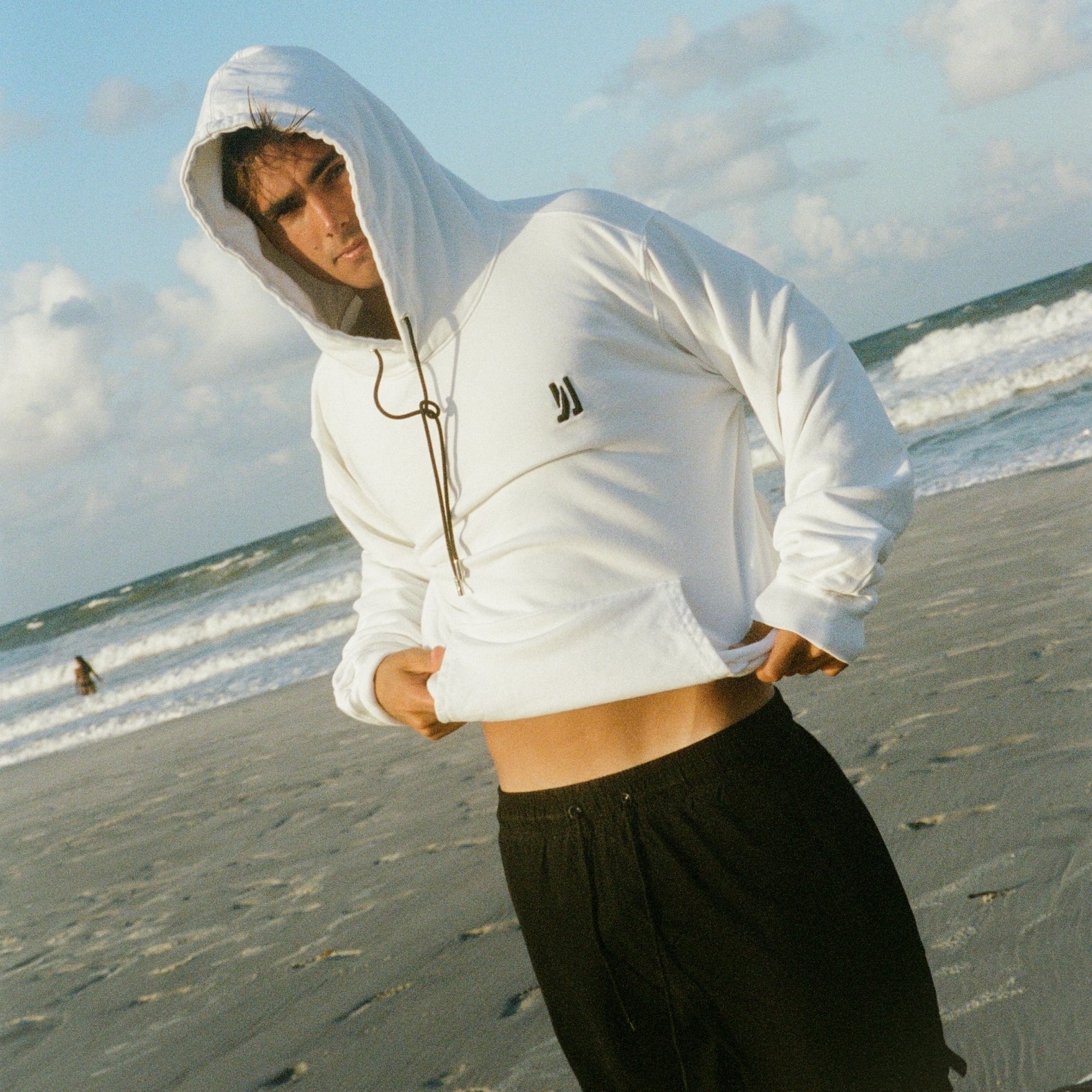 A surfer standing at the beach in front of the waves wearing Comune Luka Hoodie. COMUNE LUKA HOODIE | White