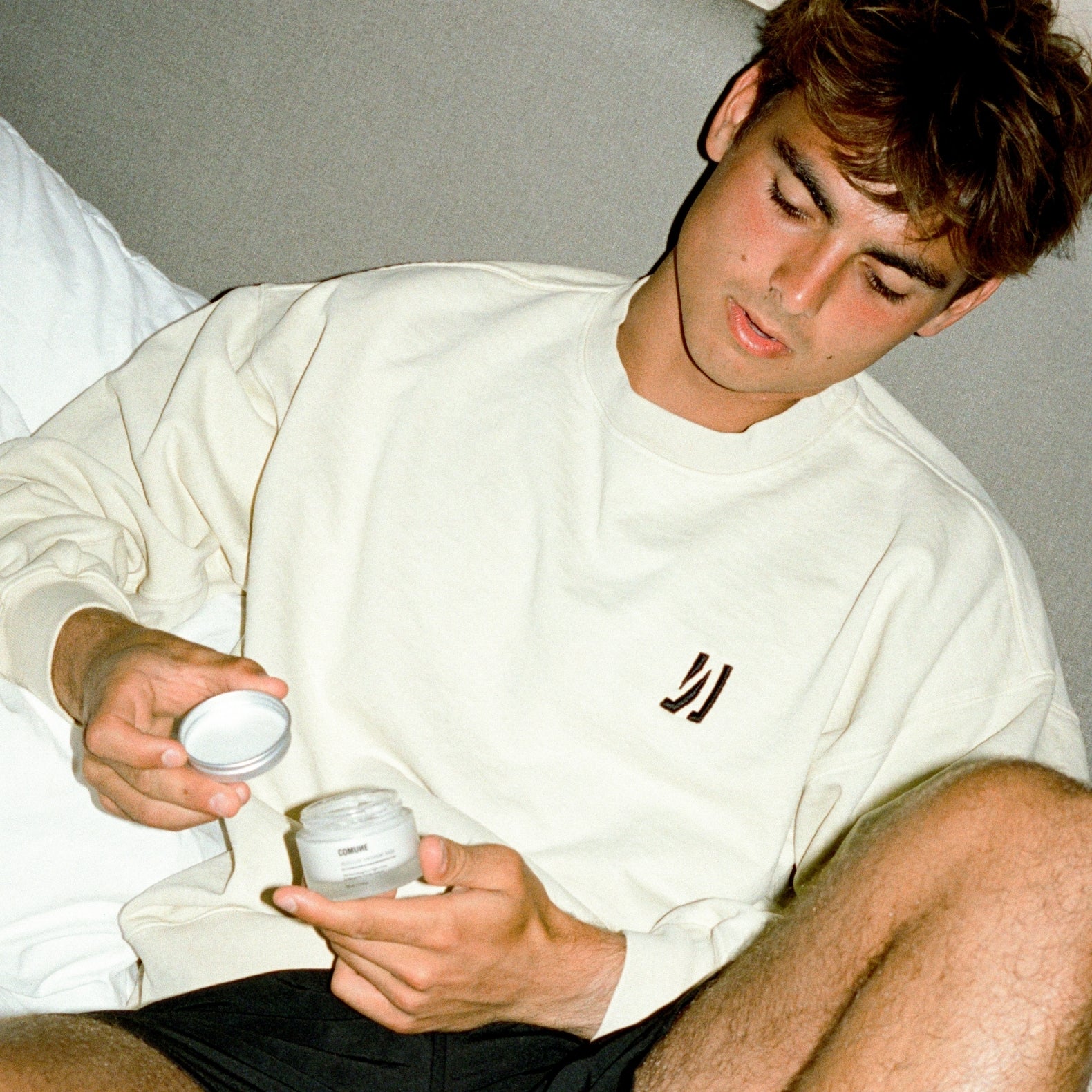 Man wearing the COMUNE Duke Crew heavyweight cotton sweatshirt in bone color holding a jar of COMUNE Revitalize Overnight Mask on a neutral background.