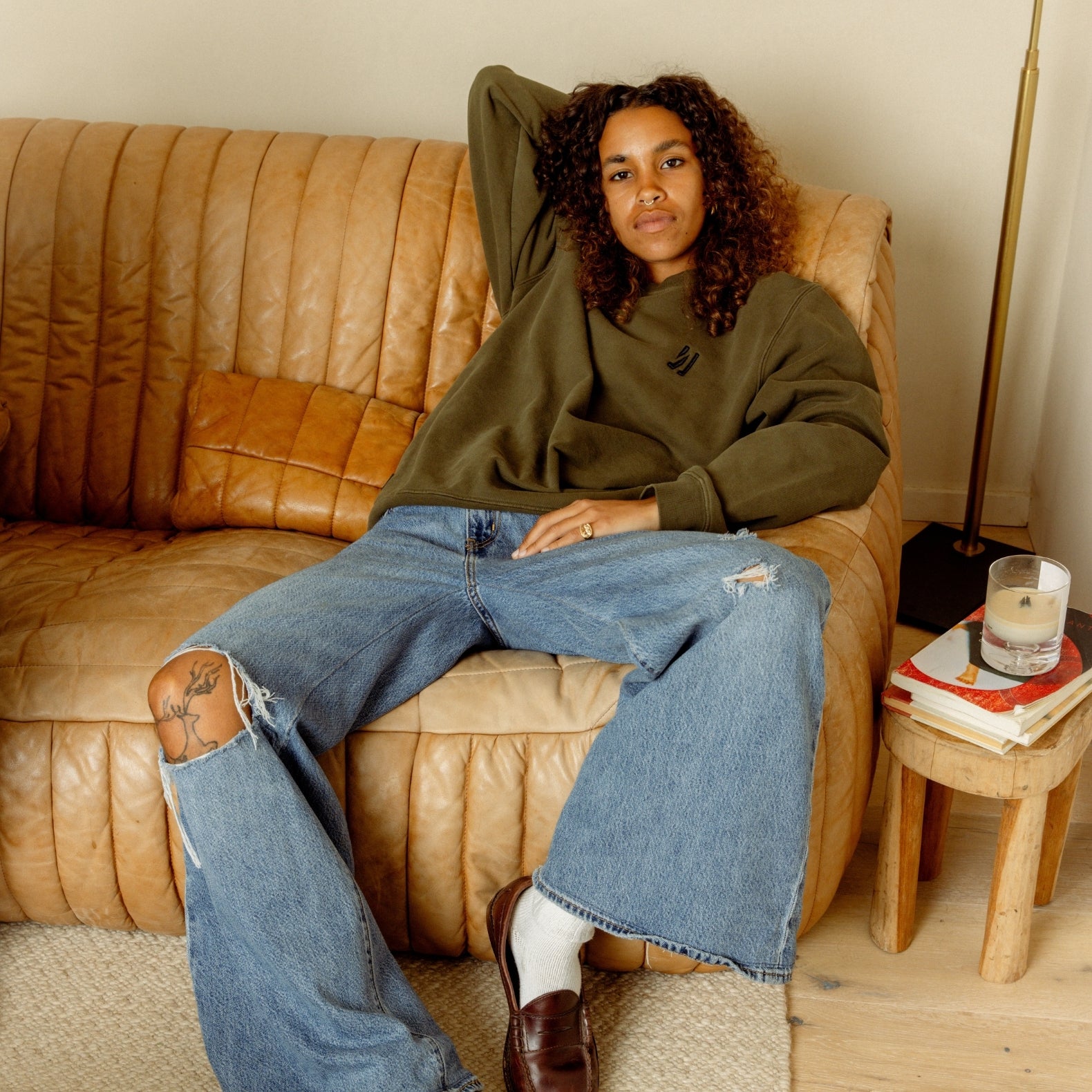 A women sitting on a brown couch wearing an army green Luka Crew with beige background. COMUNE Luka Crew | Army Green