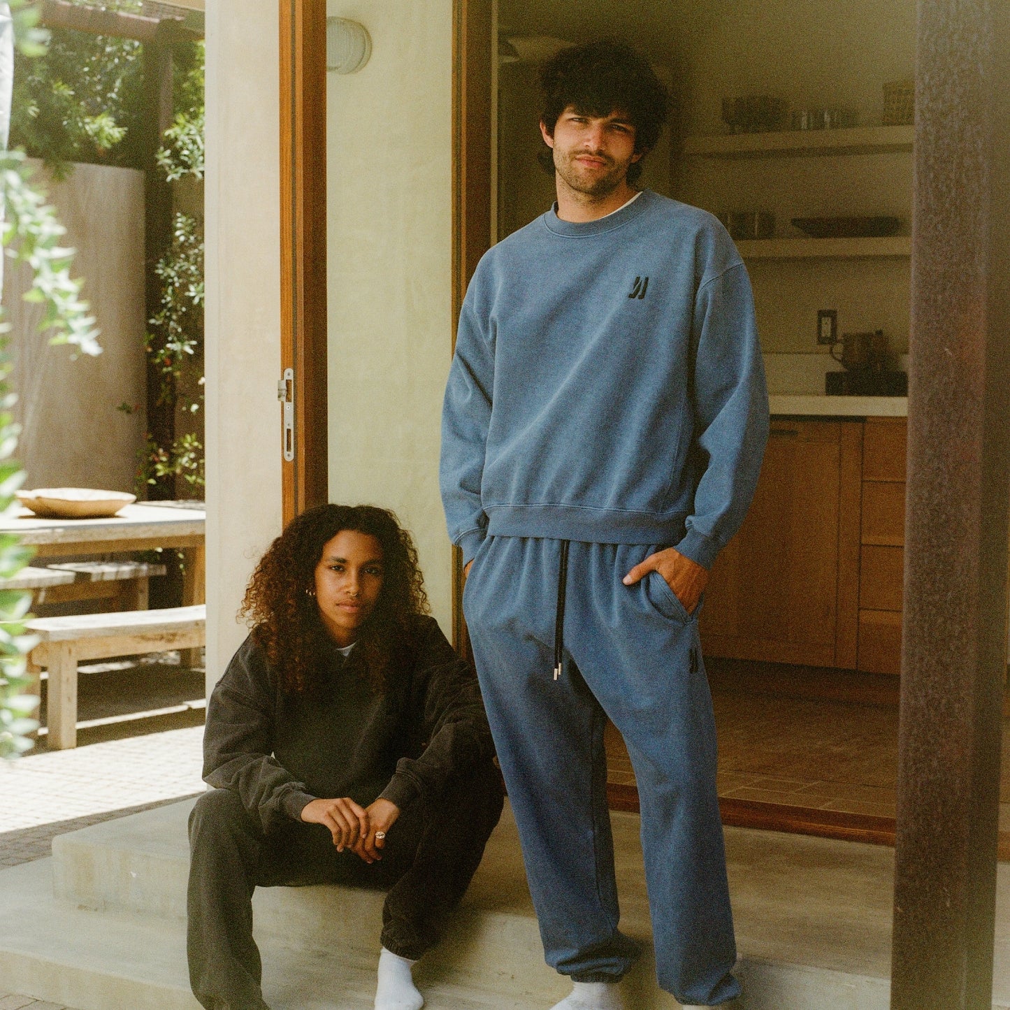 Two people in matching blue tracksuits standing in a modern kitchen, women wearing Duke hoodie and Enzo joggers in faded black color, and man wearing Duke crew and Enzo joggers in marine color. COMUNE Enzo Jogger | Marine