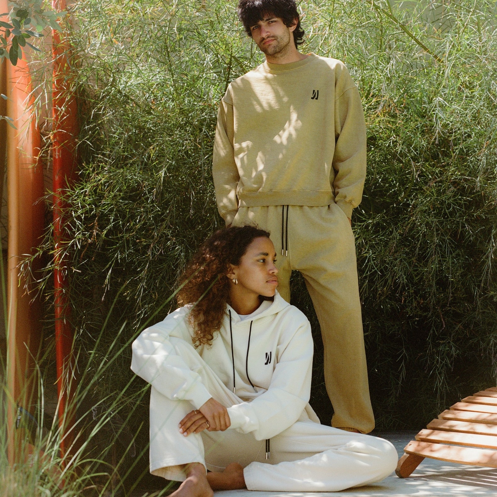 Two people wearing matching COMUNE tracksuits in beige and white, where the women is seated and the man is standing outdoors with green foliage background.