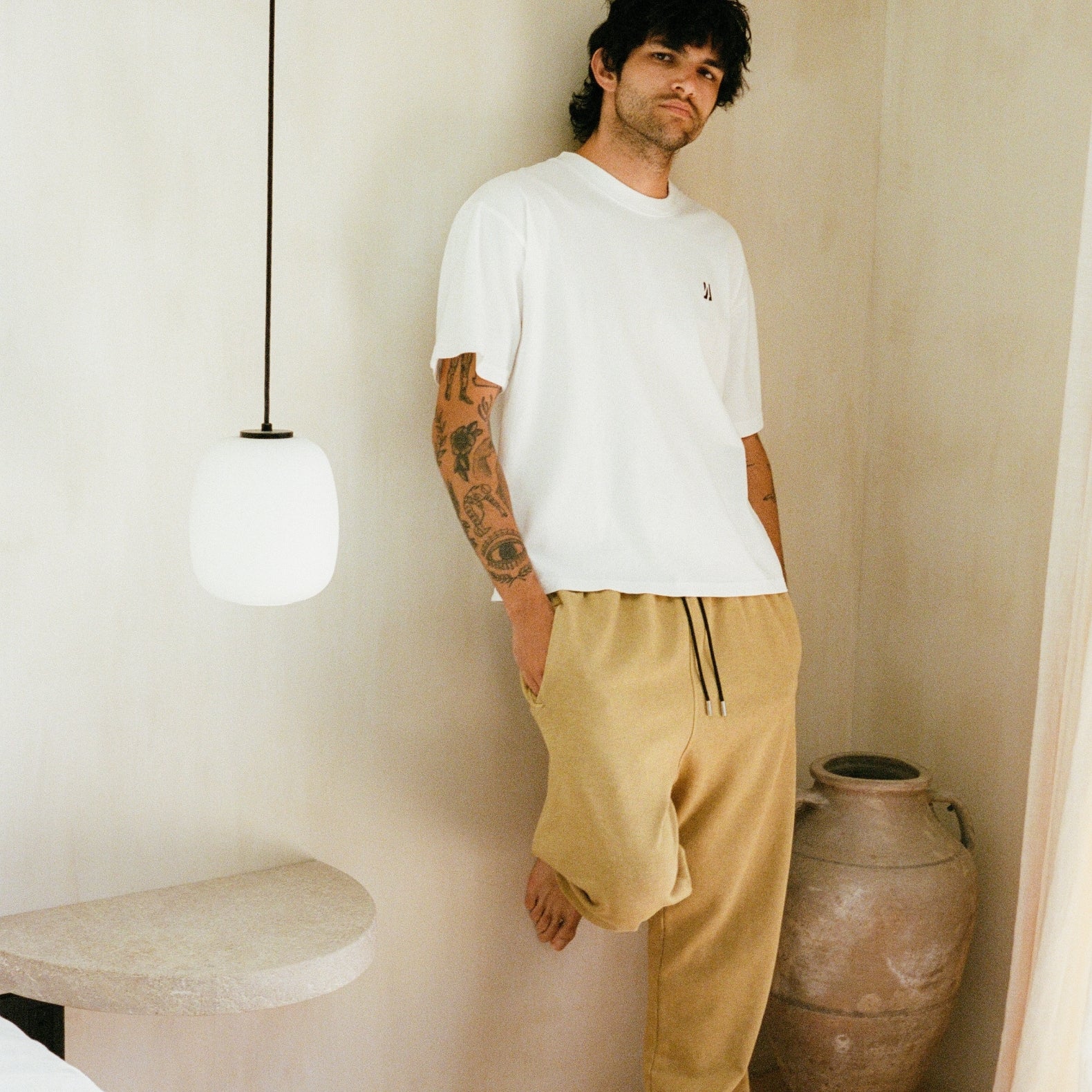Person standing and leaning against a natural color background wearing COMUNE Ryder oversized tee in white color and Enzo joggers in acorn color. COMUNE Nori Tee | White