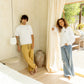 Man standing and leaning on a natural color wall wearing white Ryder oversized tee and acorn Enzo Joggers next to a women wearing Luka crew in white with jeans in front of the backyard door. COMUNE Nori Tee | White