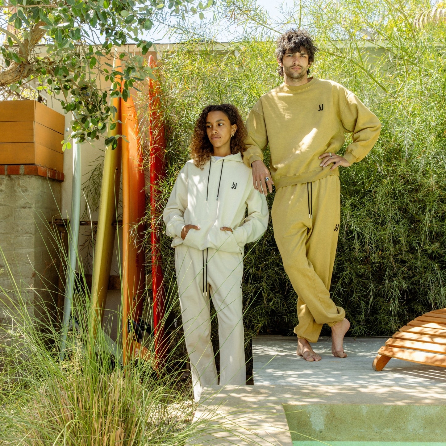 Two people standing backyard by the pool infront of greenery with 4 tall surfboard in the background, women wearing bone color Luka Crew and Duke Sweatpants, and men wearing Acorn color Duke Crew and Enzo sweatpants. COMUNE Duke Crew | Acorn