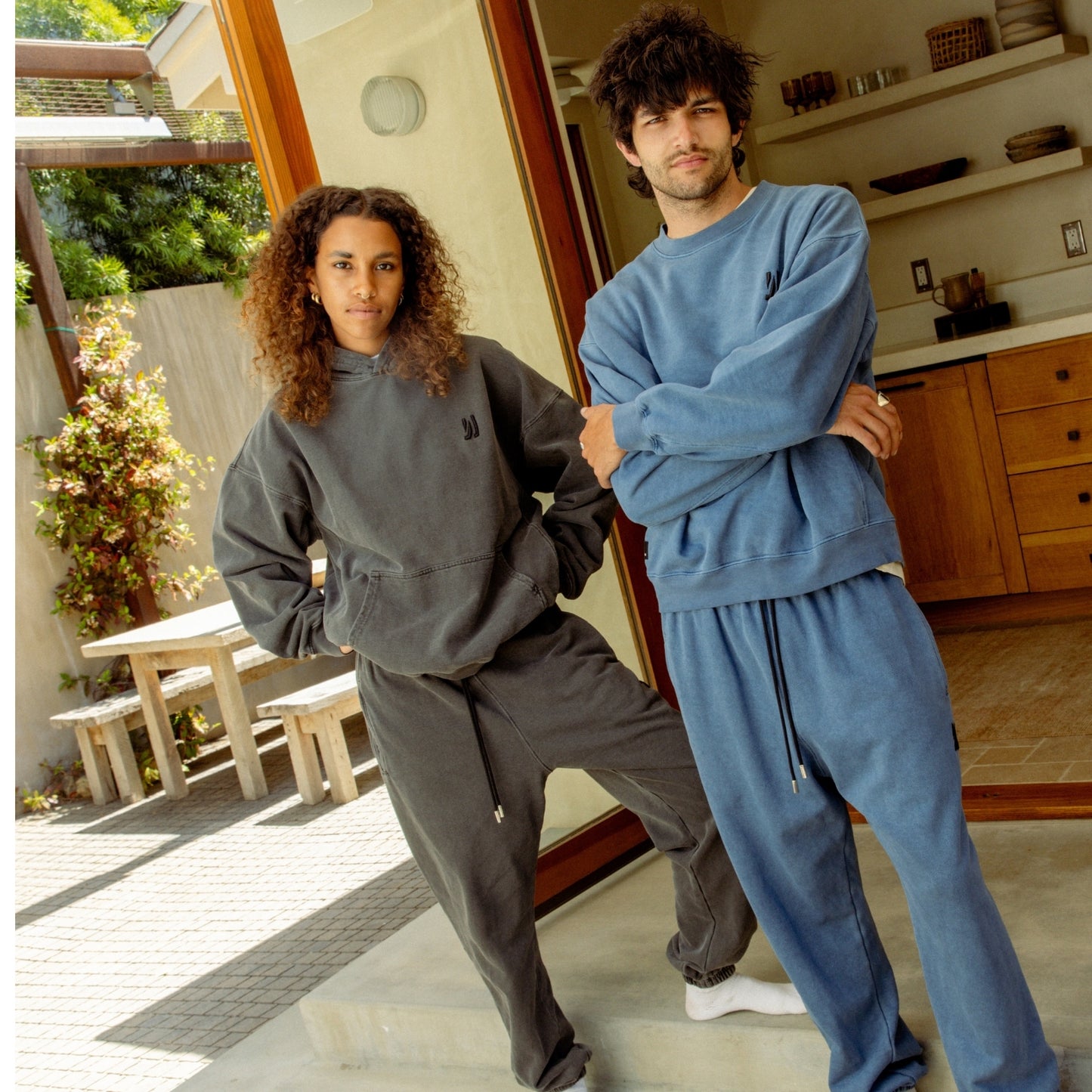 Two people standing in front of an open kitchen leading to the patio, women waring Duke Hoodie and Enzo Jogger in color faded balck, and men wearing Duke Crew and Enzo Jogger in marine color. COMUNE Duke Hoodie | Faded Black