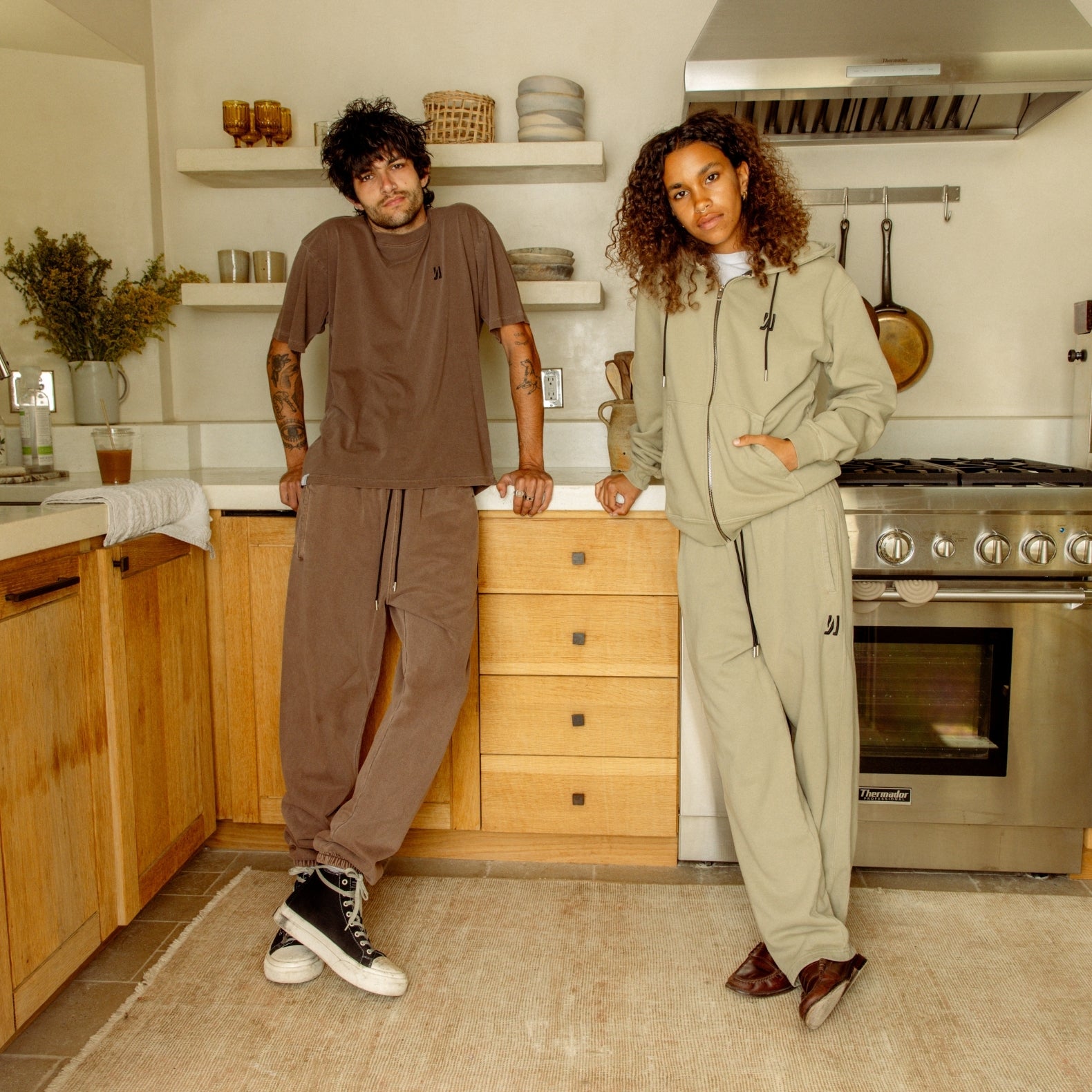Two people standing in a kitchen wearing coordinated outfits, man wearing Ryder Tee and Enzo Jogger in brown color, and women wearing Luka Zip-up hoodie and Duke sweatpants in sage color. COMUNE Enzo Jogger | Brown