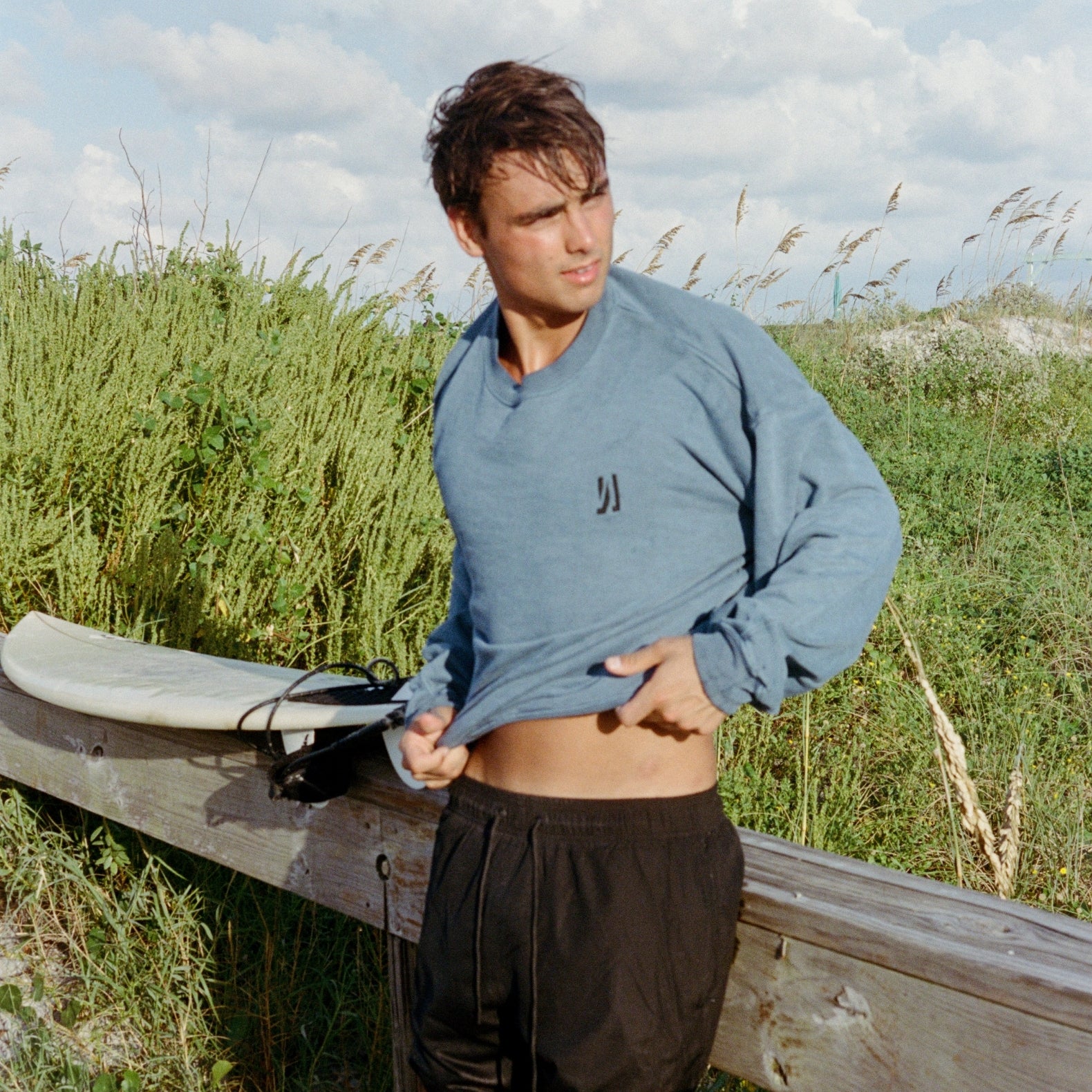 A persong leaning on a wooden deck wearing Comune Duke Crew in heavyweight cotton sweatshirt in marine color, greenery and sand background. COMUNE Duke Crew | Marine