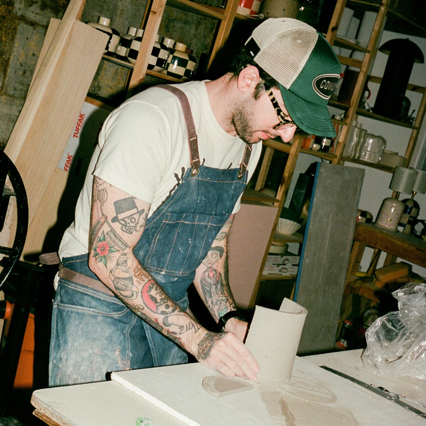 COMUNE artist Ethan wearing overalls and a Comune logo patch cap in a workshop setting.  