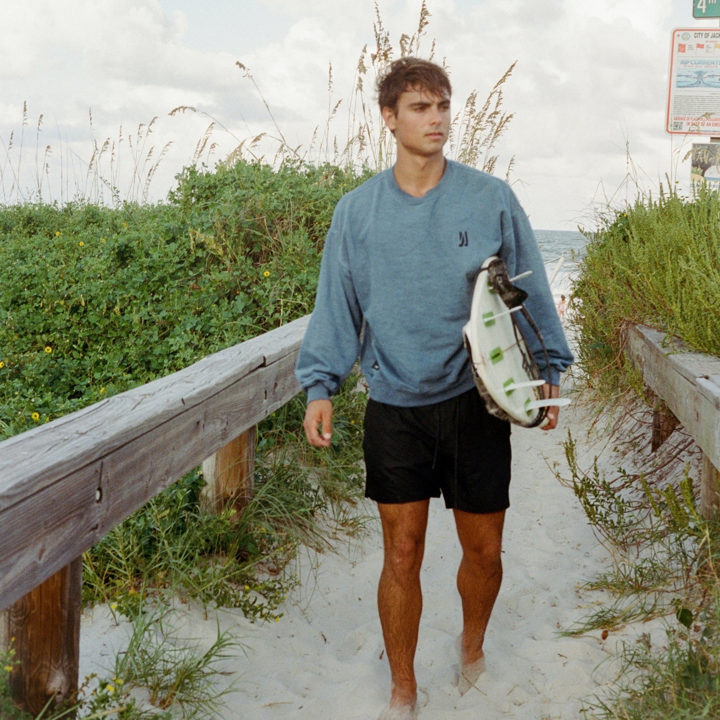 A surfer walking down on a sandy path at the beach holding his surfboard wearing Comune Duke Sweatshirt in marine color. COMUNE Duke Crew | Marine