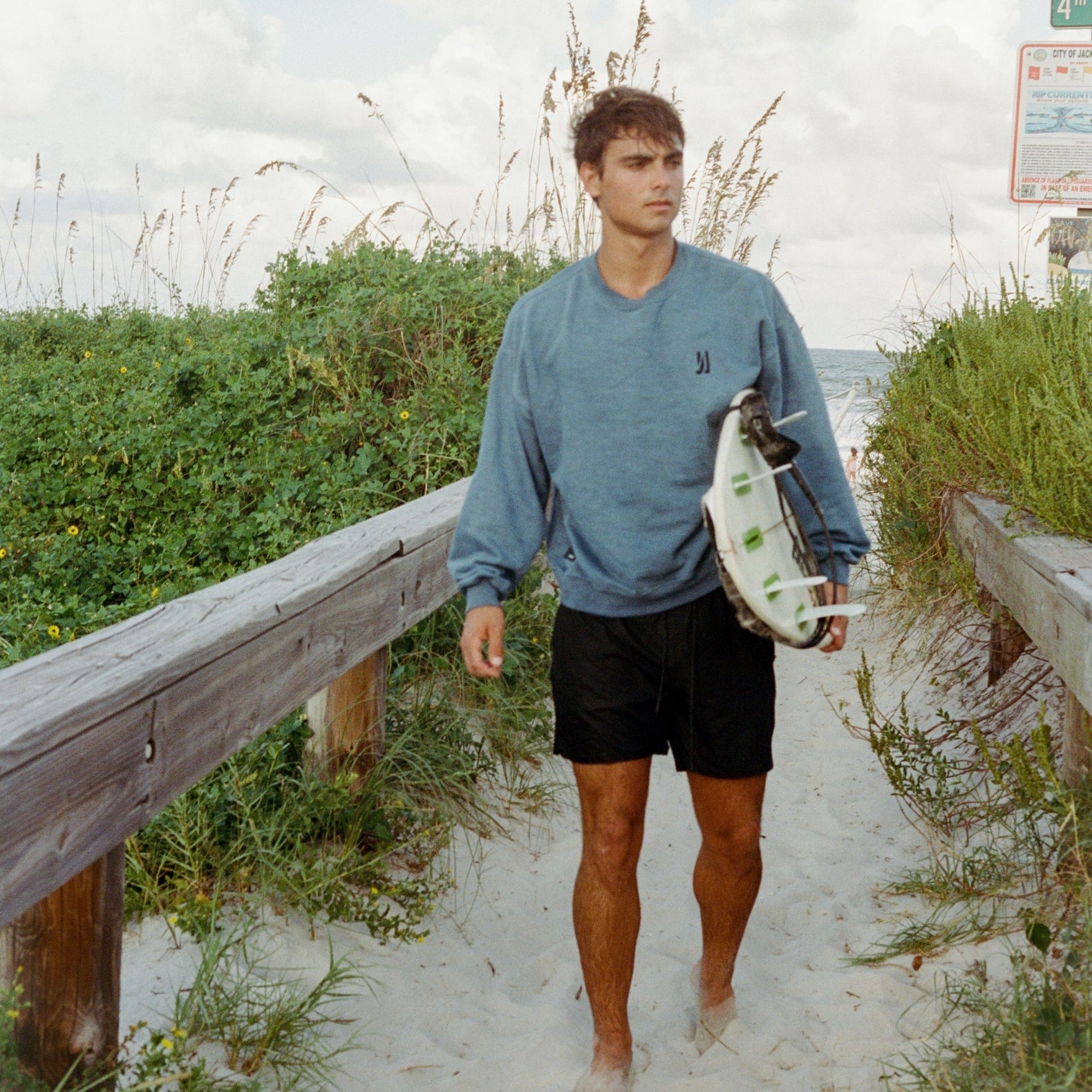A surfer walking down on a sandy path at the beach holding his surfboard wearing Comune Duke Sweatshirt in marine color. COMUNE Duke Crew | Marine