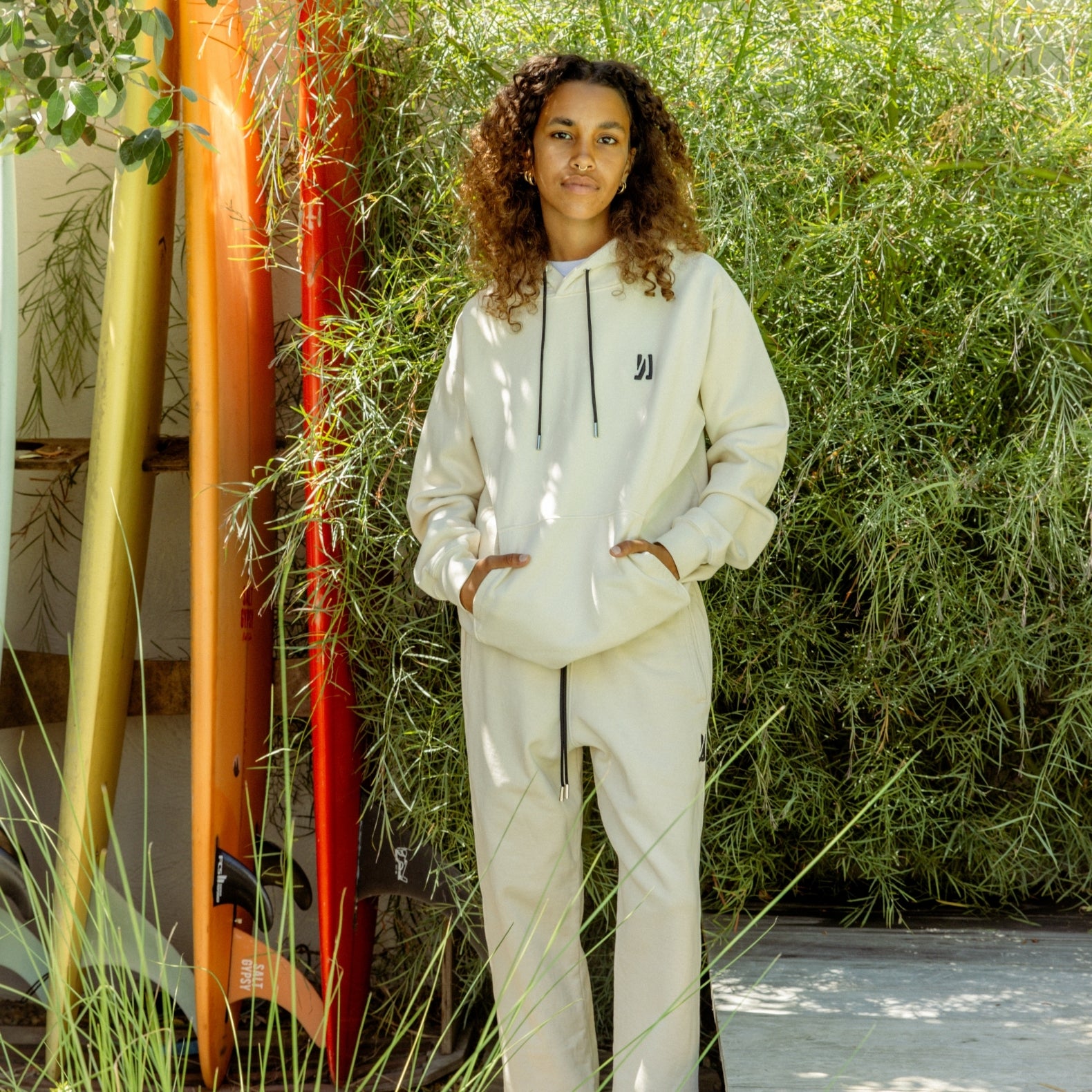 Person wearing Comune Luka Hoodie and Duke sweatpants outdoor in front of a greenery background and several surfboards. COMUNE LUKA HOODIE | Bone