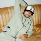 Brandon wearing a COMUNE hoodie and pants with a comune cap, sitting on a brown leather couch.