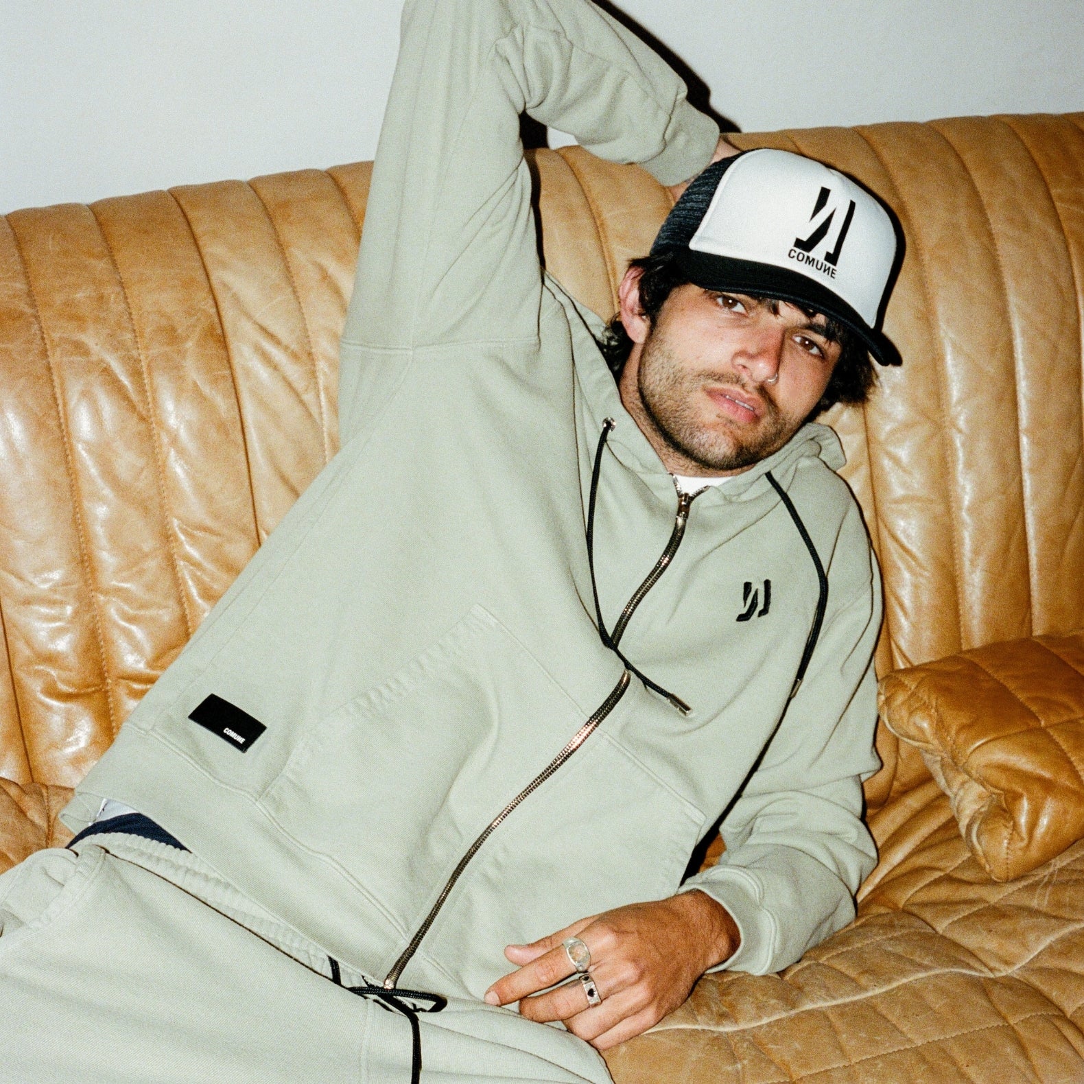 Brandon wearing a COMUNE hoodie and pants with a comune cap, sitting on a brown leather couch.