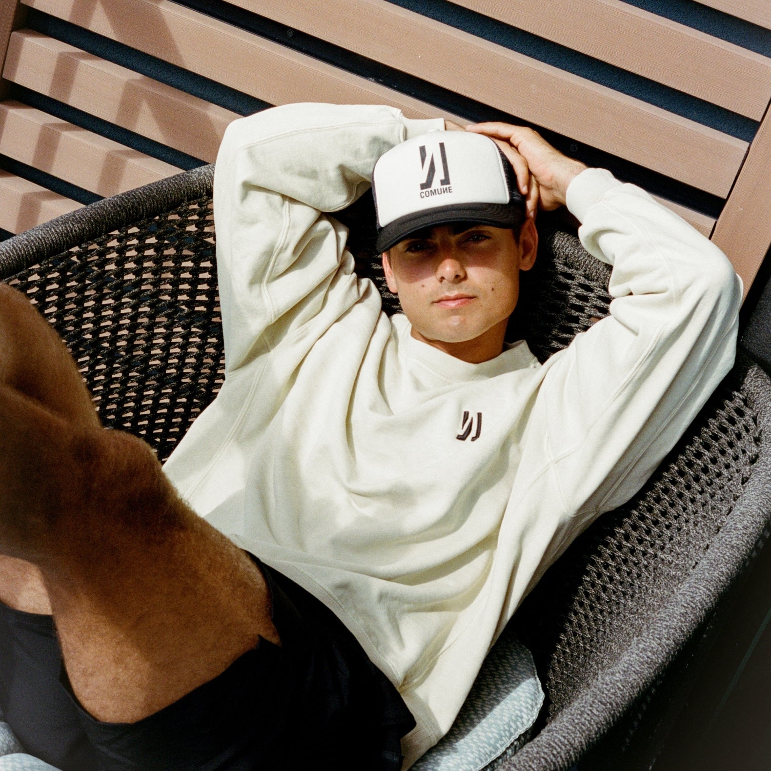 Person sitting on a gray chair backyard wearing Duke Crew sweatshirt in bone color and Icon truker hat in off-white color. COMUNE DUKE CREW | Bone