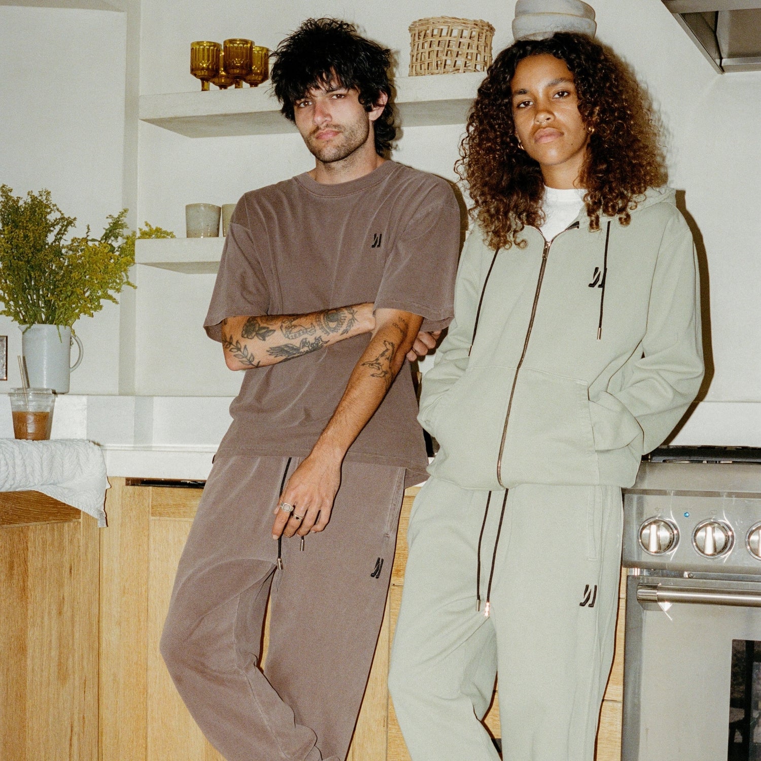 Two people standing in a kitchen wearing "COMUNE" coordinated clothing outfits.
