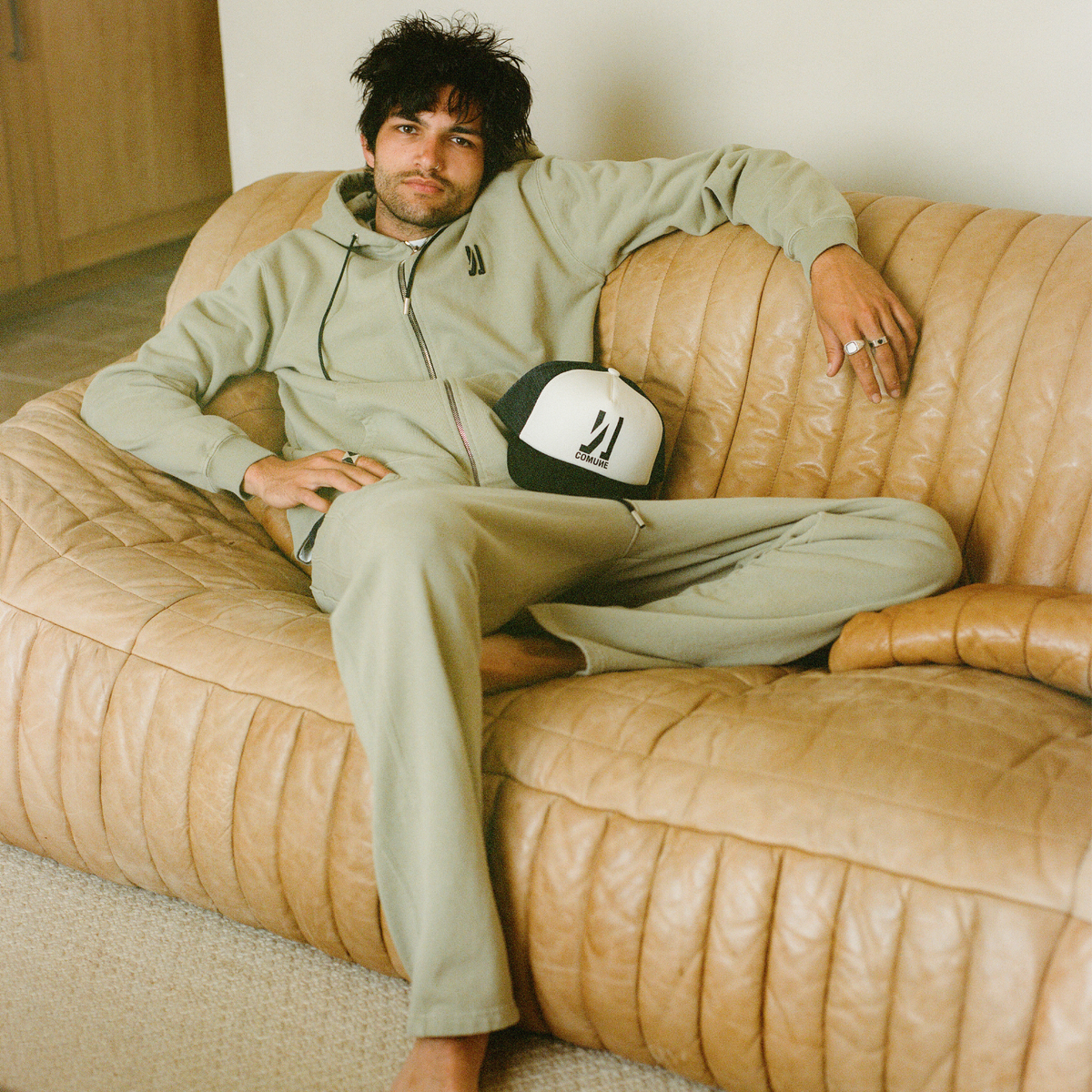 COMUNE artist Brandon sitting on a beige sofa wearing a light green COMUNE sweat  sets with a cap. COMUNE Luka Zip-up | Sage