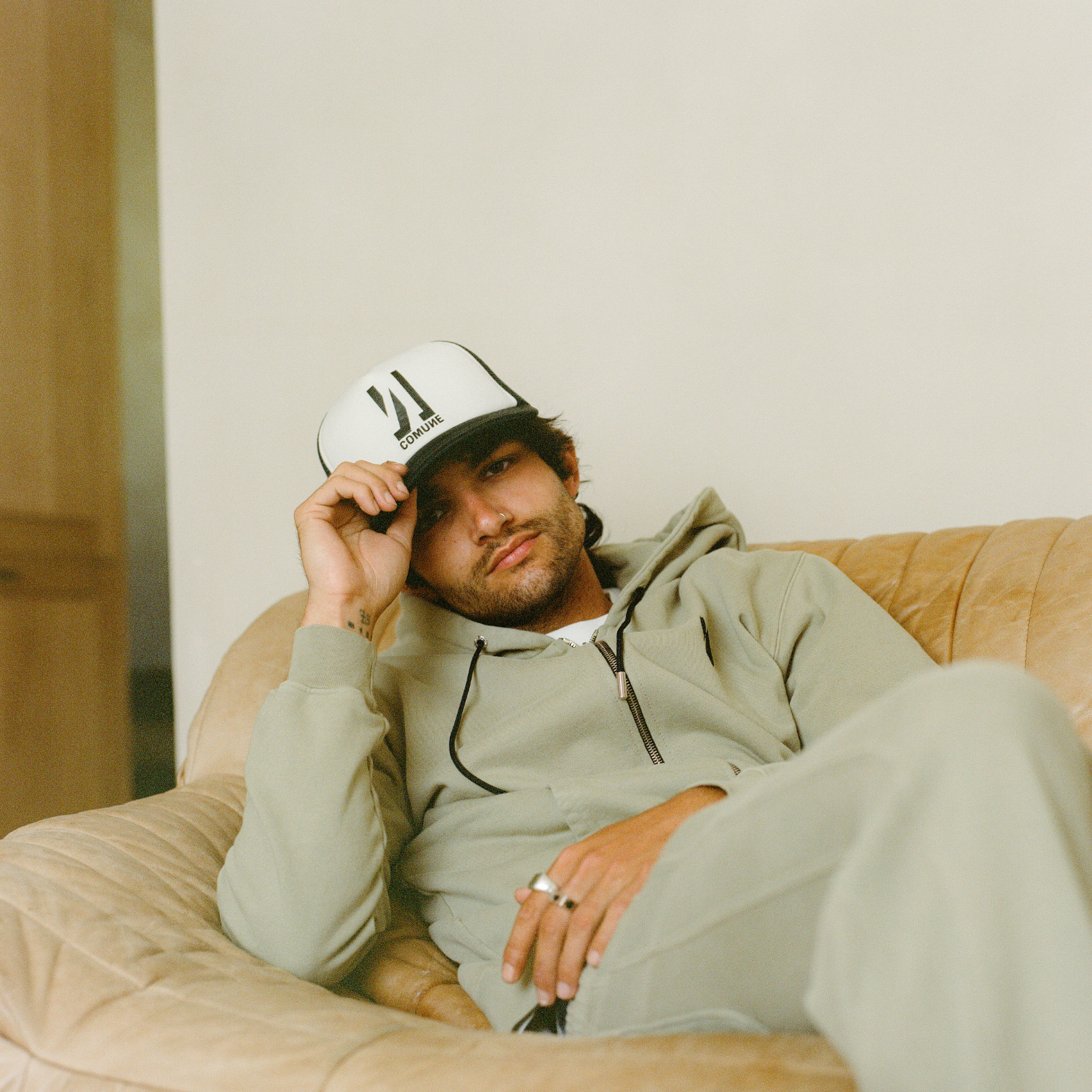 Man wearing a 'COMUNE' cap and Comune clothing, sitting on a couch in a casual setting 