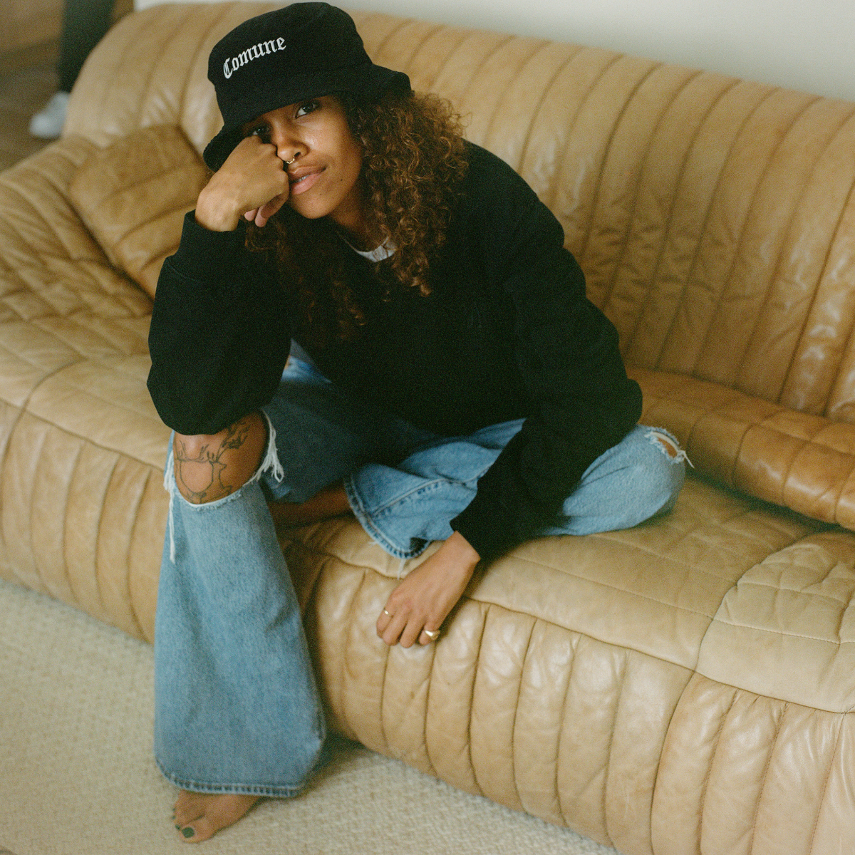 COMUNE model wearing a black COMUNE hoodie and jeans sitting on a beige sofa.