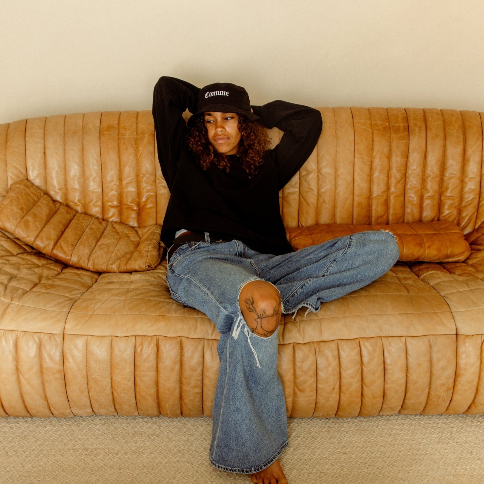 Person sitting on a brown leather couch wearing a COMUNE black sweatshirt and comune cali bucket hat