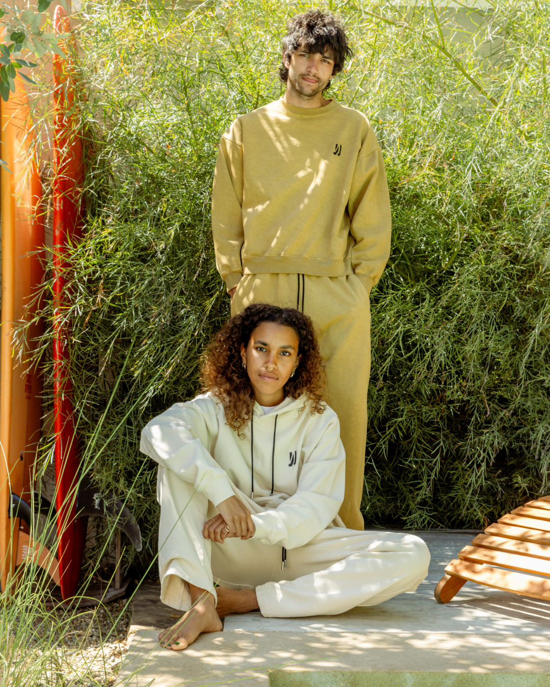 Two people wearing beige and white outfits sitting outdoors with greenery in the background