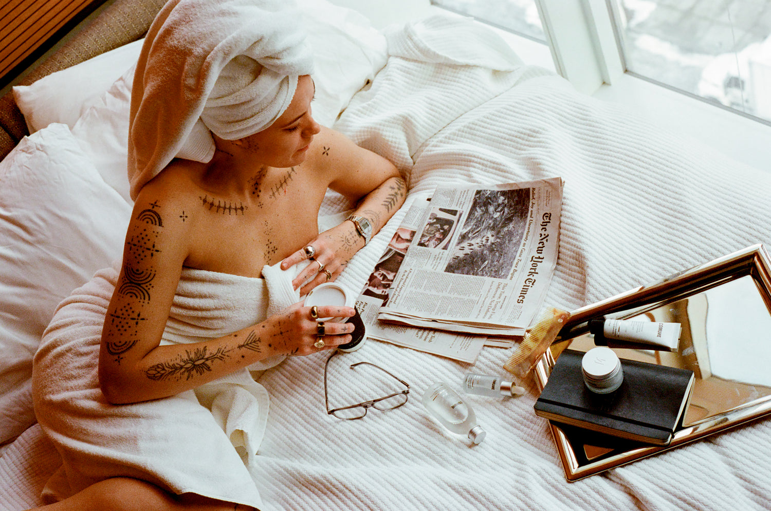 Comune model with a towel on her head reading a newspaper in bed, surrounded by personal items, including 'COMUNE' Essentials Skincare products.